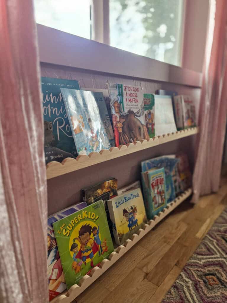 Close-up of built-in wooden picture ledges beneath a bedroom window, displaying colorful children's books framed by soft blush pink velvet curtains in a cozy girl's bedroom.