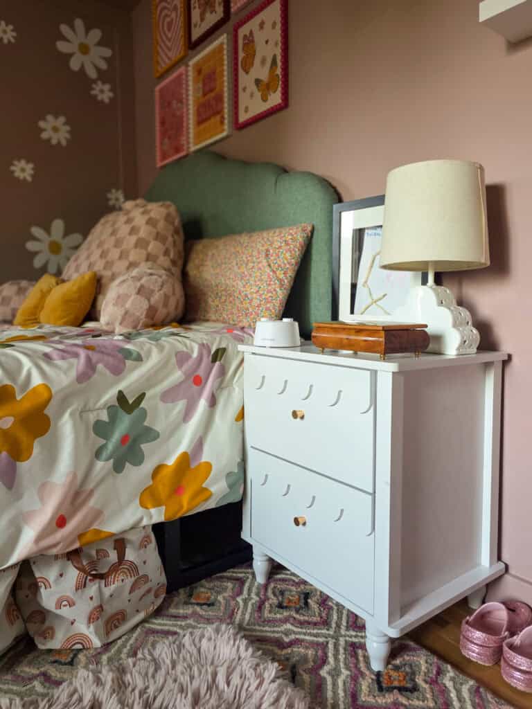 Cozy bedside view of a girl's pink bedroom featuring a white nightstand, scalloped green upholstered headboard, floral bedding, blush checkered pillows, daisy wall decals, colorful framed artwork, and a modern white table lamp.